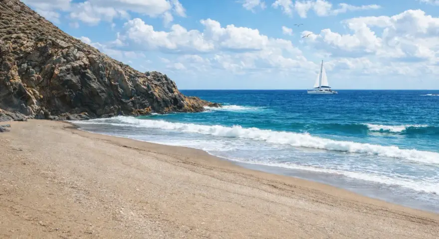 Spiaggia dorata e mare azzurro vicino a Sassari, con un catamarano bianco che naviga all'orizzonte sotto un cielo nuvoloso.