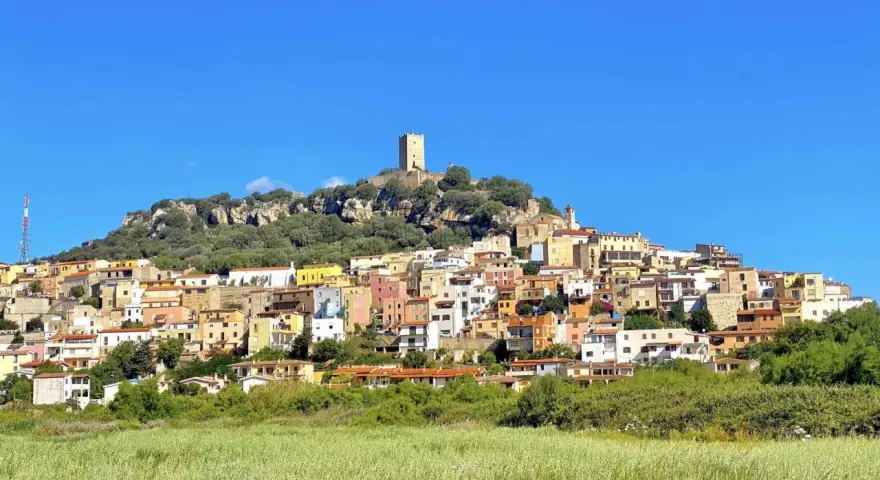 La **posada di sera** si staglia su una collina rocciosa, con case colorate e una torre medievale sotto un cielo azzurro.