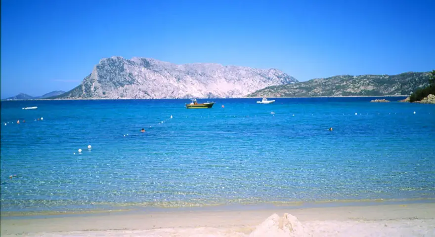 Spiaggia di Cala Suaraccia, mare cristallino, montagne sullo sfondo e barche in lontananza. Come arrivare: goditi il panorama!