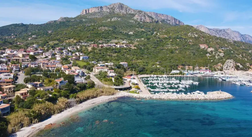 Vista aerea del centro storico di Santa Maria Navarrese, con case arroccate sulla collina, un porto turistico pieno di barche e il mare cristallino.