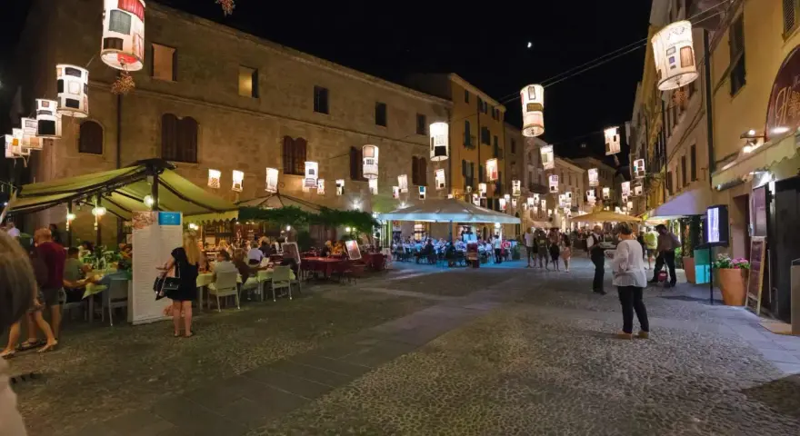 Serata ad Alghero: ristoranti all'aperto illuminati da lanterne, gente che passeggia e cena.