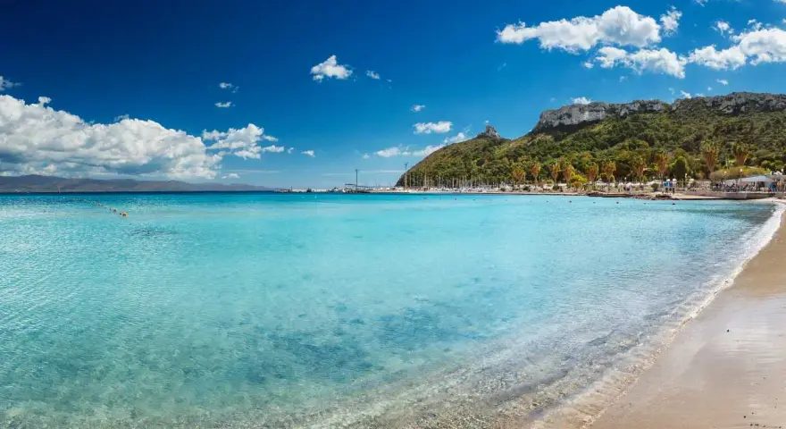 Acque cristalline e sabbia dorata sulle spiagge di Cagliari, con il promontorio verdeggiante e il porto turistico sullo sfondo.