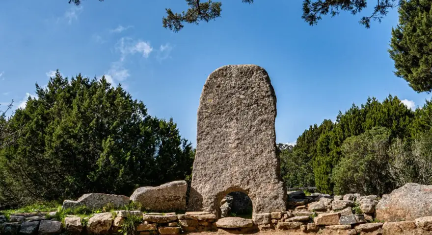 La tomba dei giganti in Sardegna, un imponente monolito di granito con un arco, circondato da pietre antiche e vegetazione rigogliosa sotto un cielo azzurro.