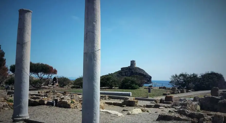 Colonne antiche nel **sito archeologico** di Nora, con una torre in lontananza su un'isola rocciosa.
