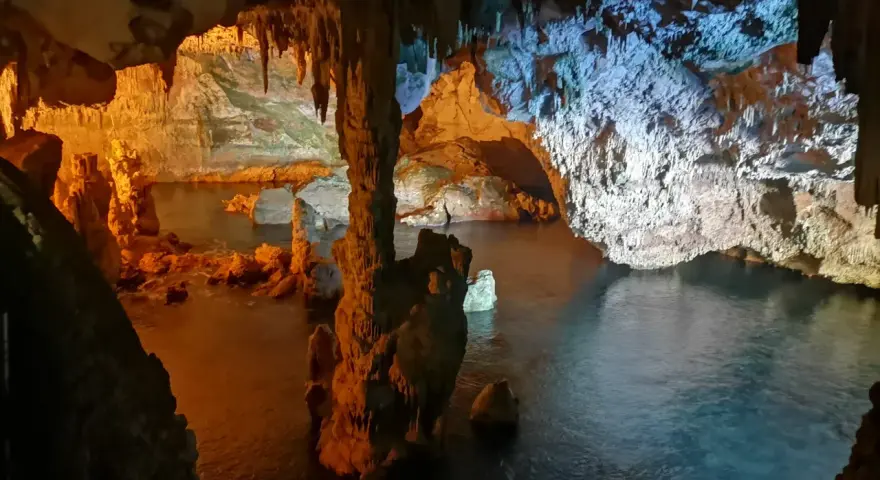 Interno di una grotta con stalattiti e stalagmiti illuminate da luci arancioni e blu. L'acqua scura riflette le formazioni rocciose in questa grotta verde.
