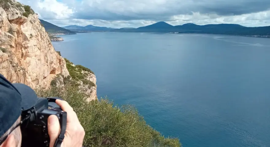 Scogliera a picco sul mare blu, con un fotografo che cattura il paesaggio. Si intravede la costa e le montagne all'orizzonte, forse vicino alla grotta verde di Alghero.