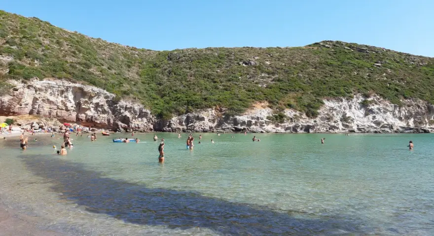 Persone si godono il sole e il mare cristallino nella spiaggia cala lunga, con colline verdi e rocce a fare da cornice.