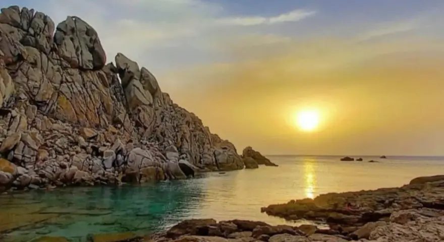 Scogliere rocciose bagnate da acque cristalline, con il sole che tramonta sulla valle della luna, creando riflessi dorati sul mare.