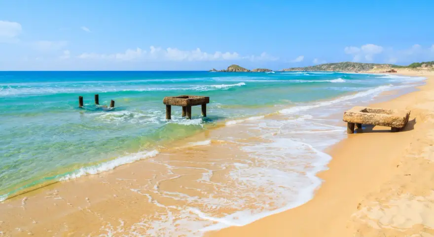Spiaggia di Chia, Cagliari. Resti di antiche strutture in mare e sulla sabbia dorata, con acque turchesi e cielo azzurro.