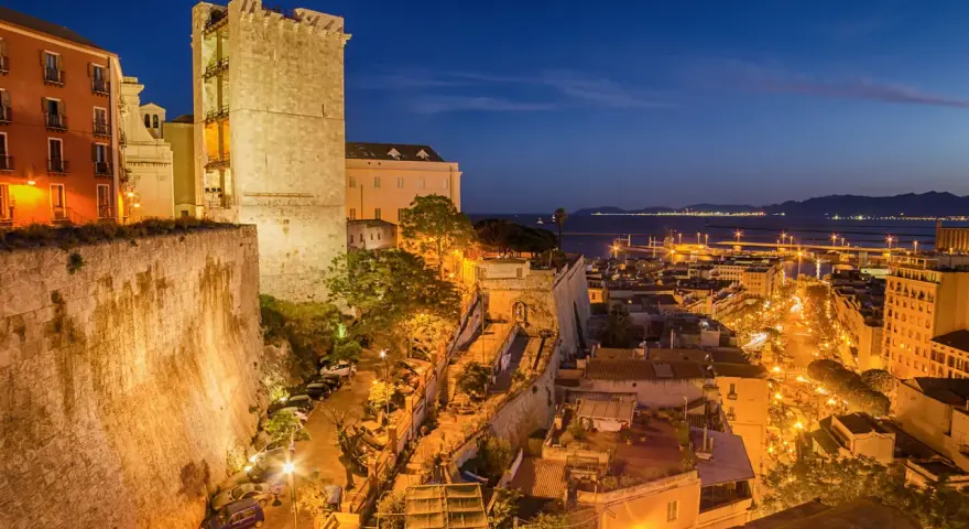 Torre medievale e palazzi illuminati nella piazza di Cagliari, con il porto sullo sfondo al crepuscolo.