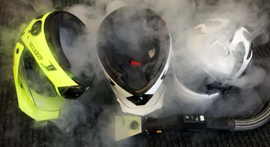 Tre caschi, uno giallo fluo, uno bianco e nero, e uno bianco, sono avvolti dal vapore. Un accessorio per la pulizia a vapore è posizionato davanti a loro, suggerendo che il casco non deve essere pulito con solventi.