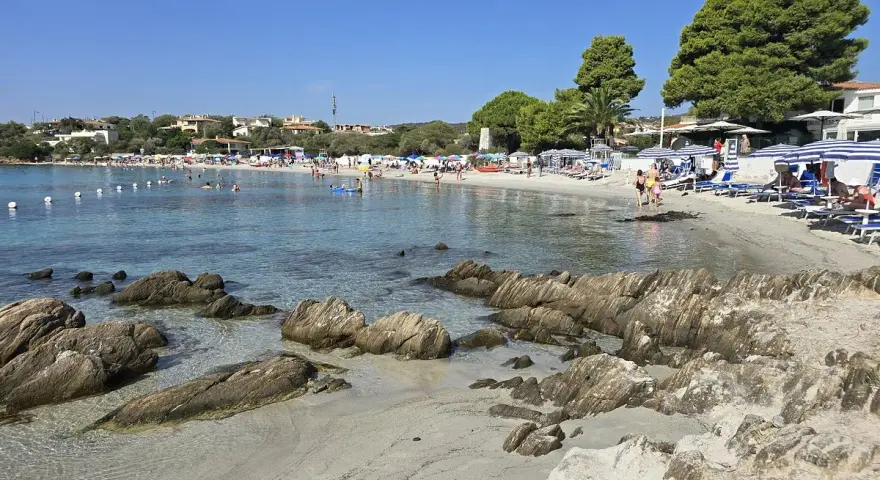 Spiaggia del Pellicano: sabbia chiara, rocce levigate e mare cristallino con bagnanti e ombrelloni.