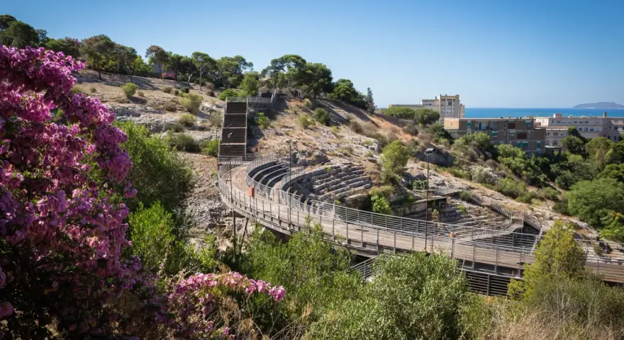 Un suggestivo anfiteatro a Cagliari, con gradinate in pietra e passerelle in legno, immerso nel verde e con fiori viola in primo piano.