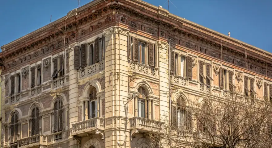 Angolo del **Palazzo Accardo a Cagliari**, con facciata in pietra chiara, balconi in ferro battuto e finestre con persiane.