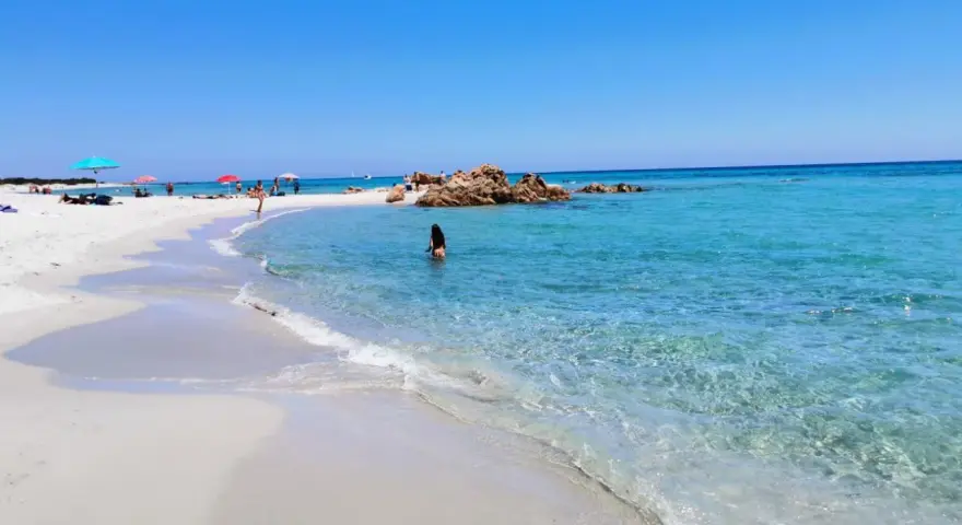 Spiaggia di Berchida: sabbia bianca, acque cristalline e rocce rosse sotto un cielo azzurro. Una donna si gode il mare calmo.