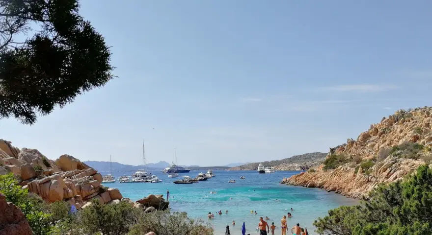 Spiaggia affollata a Cala Coticcio, con gente che prende il sole e nuota nelle acque cristalline. Diverse barche e yacht ancorati nella baia.