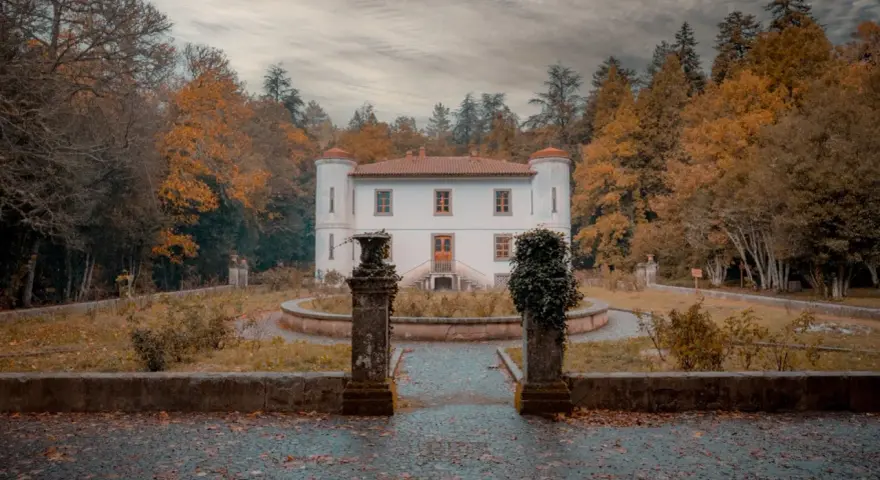 Un'antica villa bianca con torrette, circondata da alberi autunnali dai colori caldi. Un sentiero lastricato conduce all'ingresso, tra statue e vegetazione. Le **badde salighes** sussurrano storie di tempi passati.