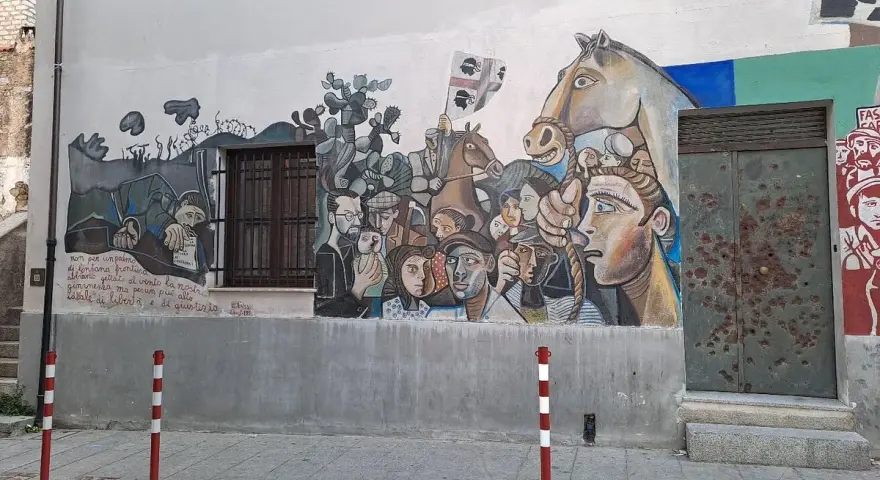 Un grande murale colorato decora un edificio in un paese, con volti, un cavallo e bandiere. Un'opera d'arte che racconta storie di un paese, murales in Sardegna.