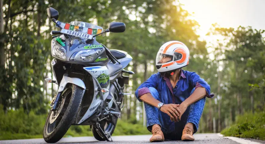 Giovane con casco da moto arancione e bianco, seduto accanto alla sua Yamaha. Spera di allargare il casco stretto per un viaggio più comodo.