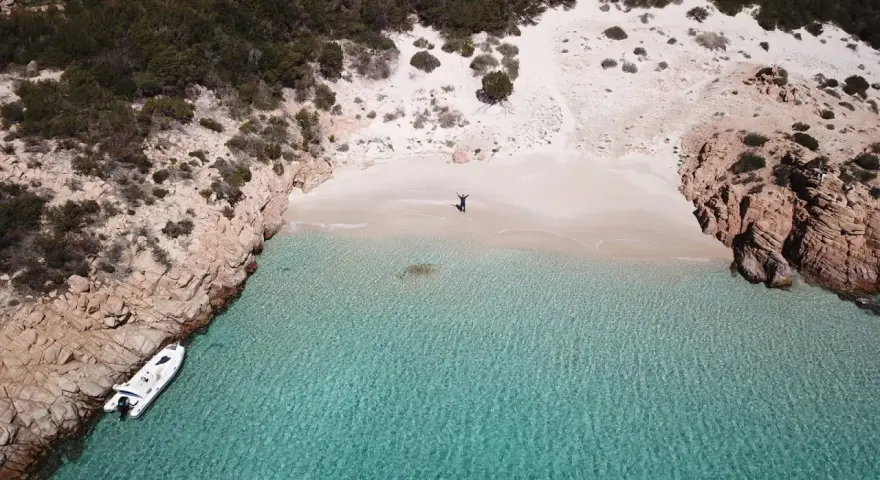 Una persona celebra la bellezza di Cala Soraya, con acque cristalline e rocce rosse. Una barca è ormeggiata.