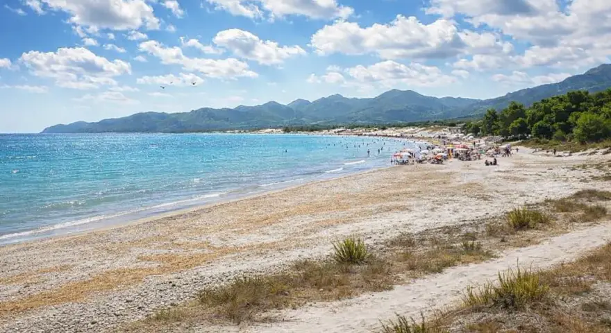 Spiaggia affollata con ombrelloni e gente che si gode il sole. Il nuoro mare azzurro lambisce la costa, con montagne verdi sullo sfondo.