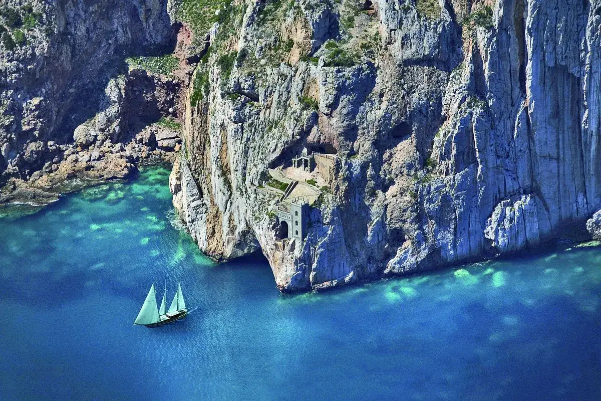 Vela solitaria naviga vicino alle scogliere di Porto Flavia, con un antico edificio incastonato nella roccia.