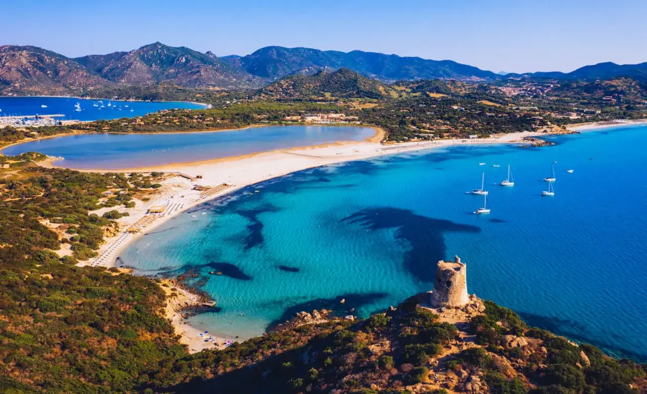 Torre costiera e acque turchesi tra le spiagge di Villasimius, con montagne sullo sfondo e barche a vela.