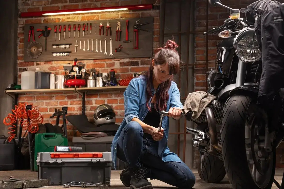 Donna al lavoro nel suo rimessaggio moto, intenta a riparare il motore con una chiave inglese. Attrezzi e moto la circondano.