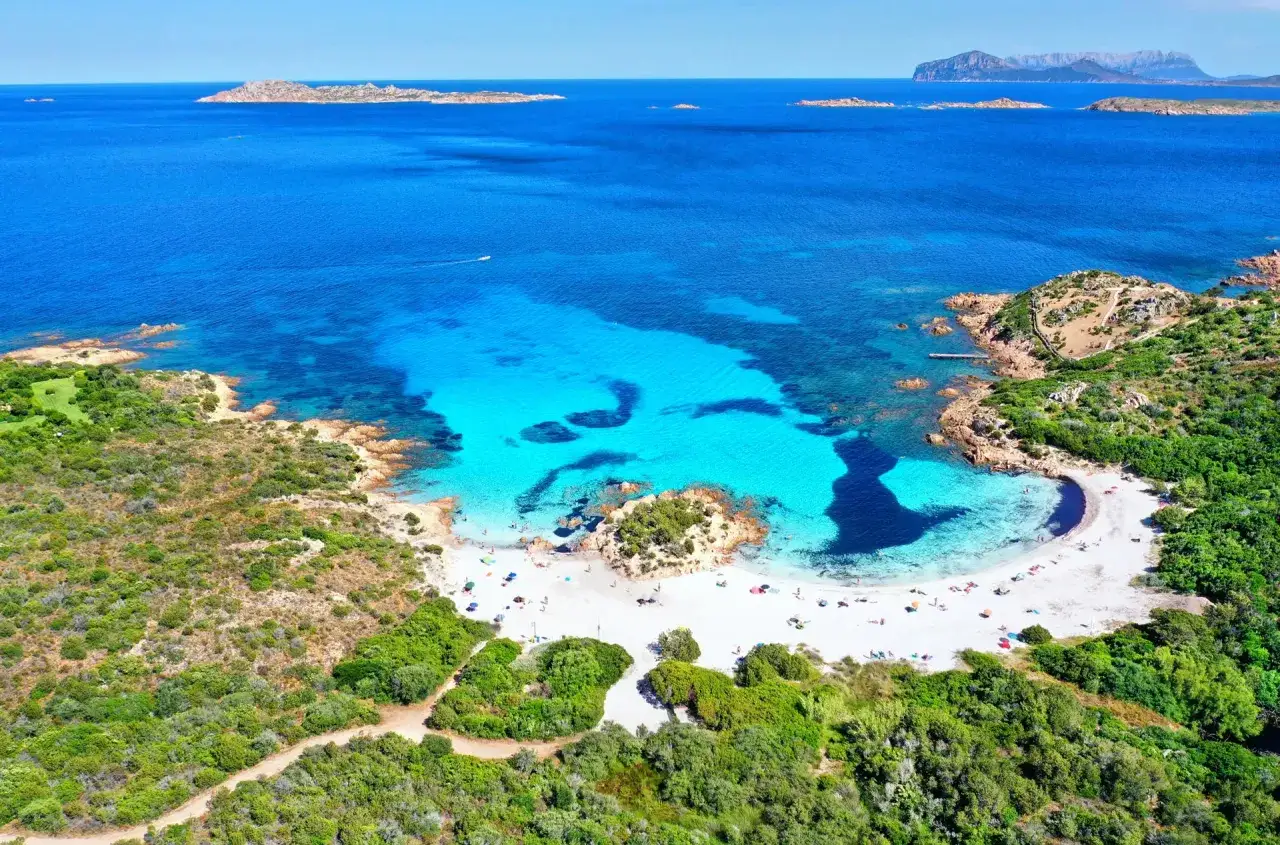 Spiaggia bianca e acque cristalline nel mare della Sardegna, con rocce e macchia mediterranea.