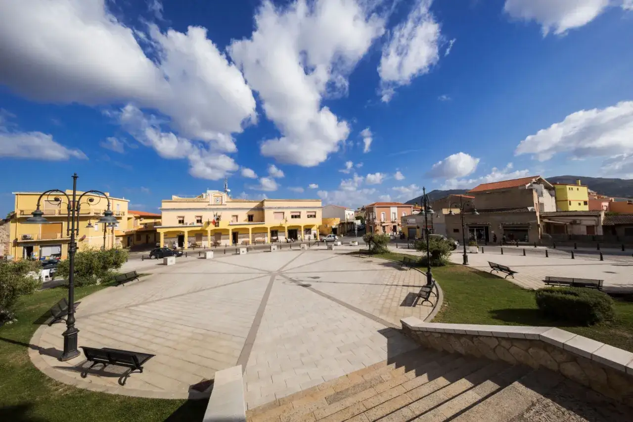 Piazza di San Giovanni Suergiu con edifici gialli e arancioni, cielo azzurro e nuvole bianche.