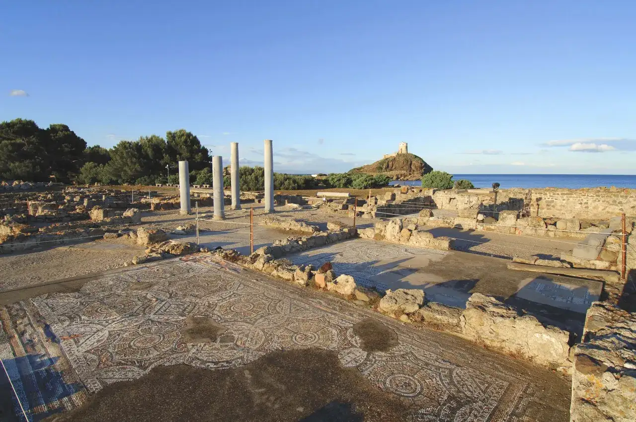 Antiche rovine del sito archeologico di Nora, con un mosaico ben conservato in primo piano e una torre su un isolotto sullo sfondo.