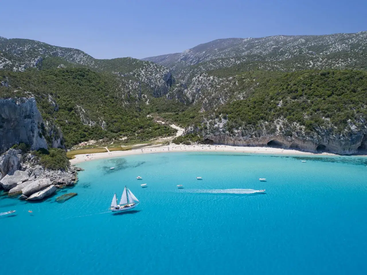 Spiaggia paradisiaca con acque cristalline e montagne verdi, un veliero solca il mare vicino a Cala Gonone.