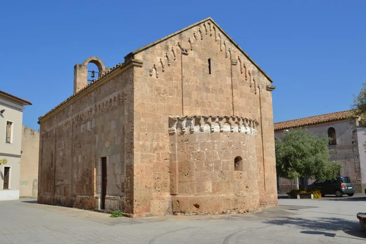 Antica chiesa in pietra con campanile a vela, decorazioni archetti e un piccolo foro. Un'auto parcheggiata e un albero completano la scena. Sa sippa.