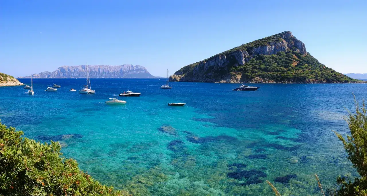 Acque cristalline e barche a vela nella splendida Cala Moresca, con la sagoma di Tavolara sullo sfondo.