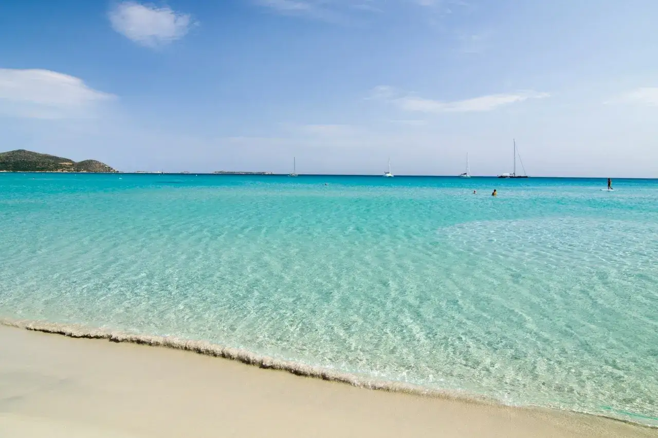 Spiaggia caraibica in Sardegna sud: cosa vedere? Acque cristalline, sabbia bianca, barche a vela e persone che nuotano e fanno SUP.