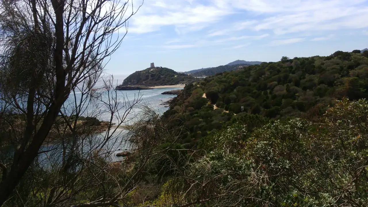Vista panoramica della costa di Chia, con la torre sul promontorio e il mare azzurro. Un sentiero si snoda tra la vegetazione rigogliosa, evocando la storia del centro storico.