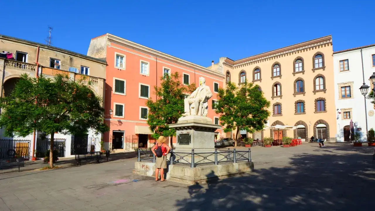 Piazza Tola a Sassari, con la statua di un uomo seduto al centro, circondata da edifici colorati e alberi verdi.