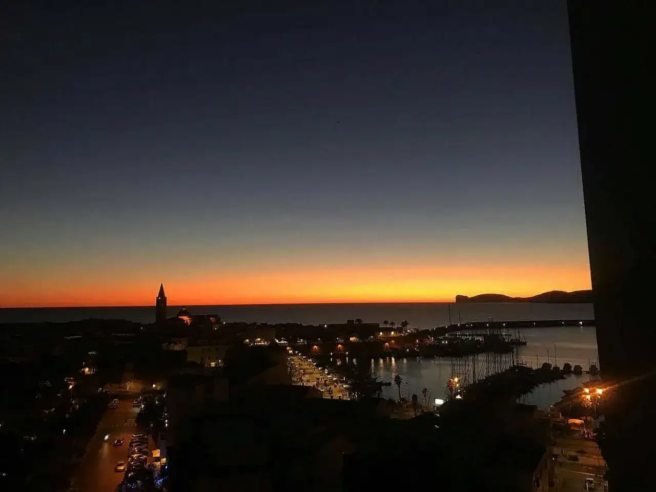 Tramonto ad Alghero, con il porto illuminato e la torre campanaria che si staglia contro il cielo arancione. Perfetto per scoprire cosa fare ad Alghero la sera.