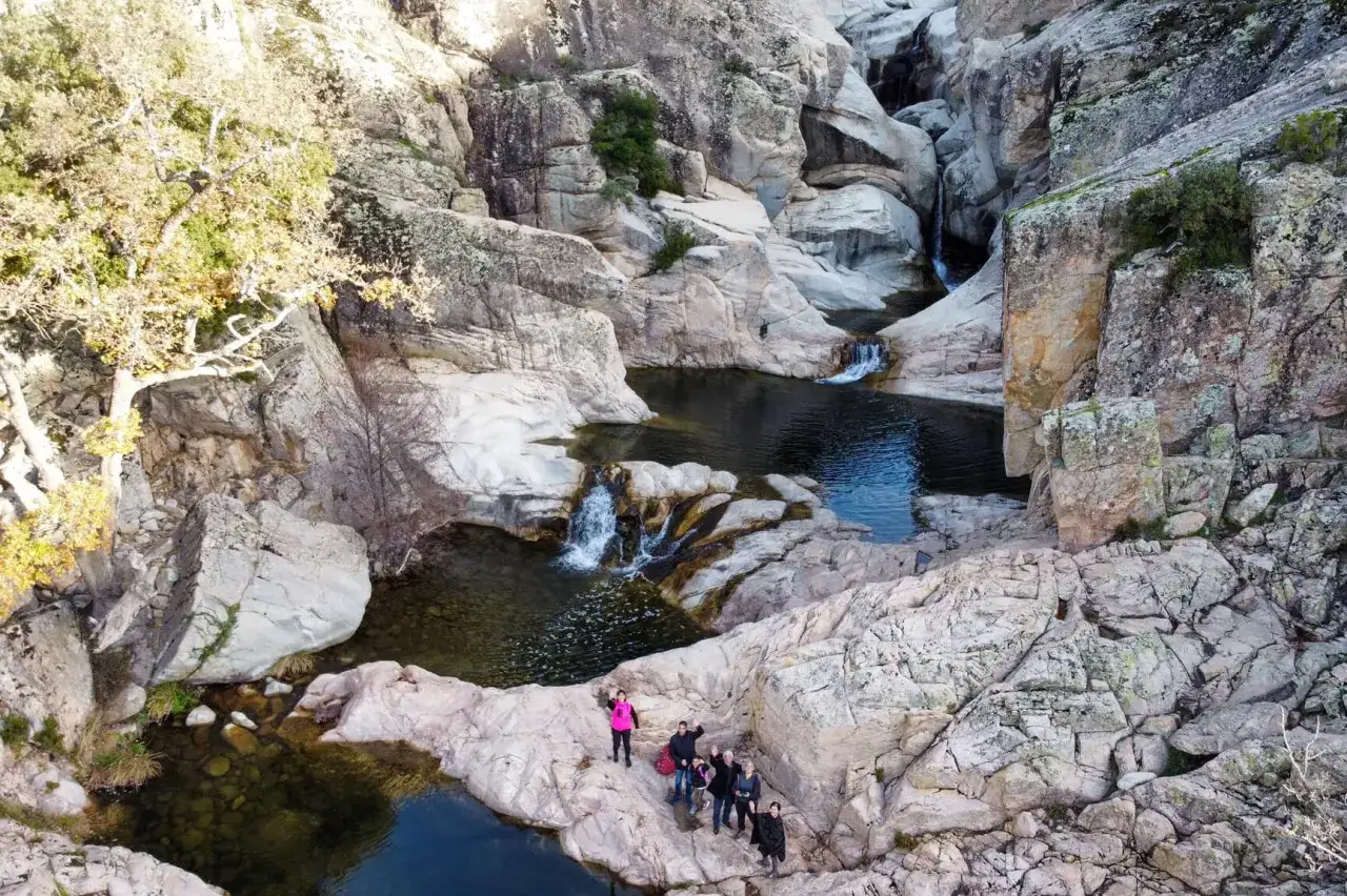 Un gruppo di escursionisti ammira un canyon con cascate e pozze d'acqua cristallina. Uno di loro, con un giubbotto rosa acceso, sembra dire