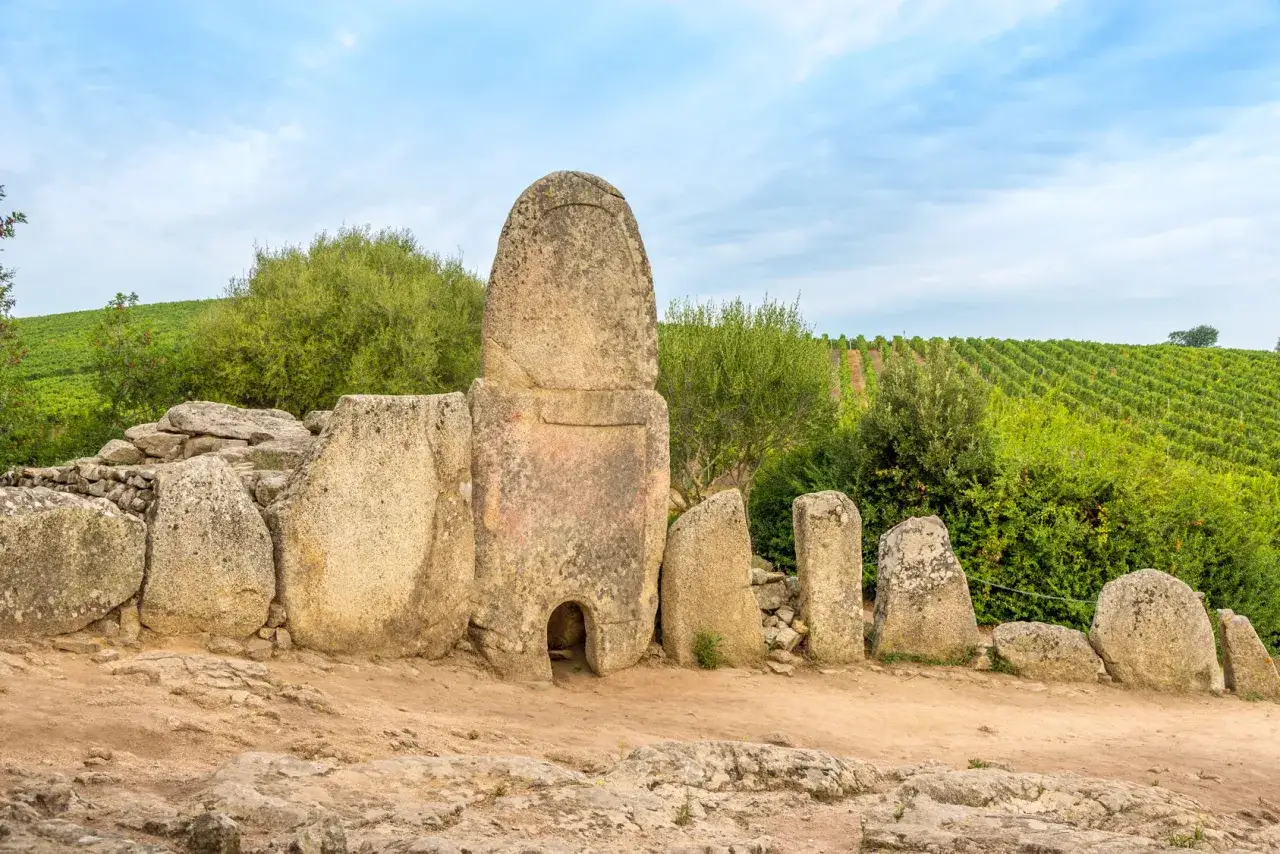 Antiche pietre di un sepolcro a corridoio, simile a un piccolo Stonehenge in Sardegna, circondate da vegetazione e vigneti.
