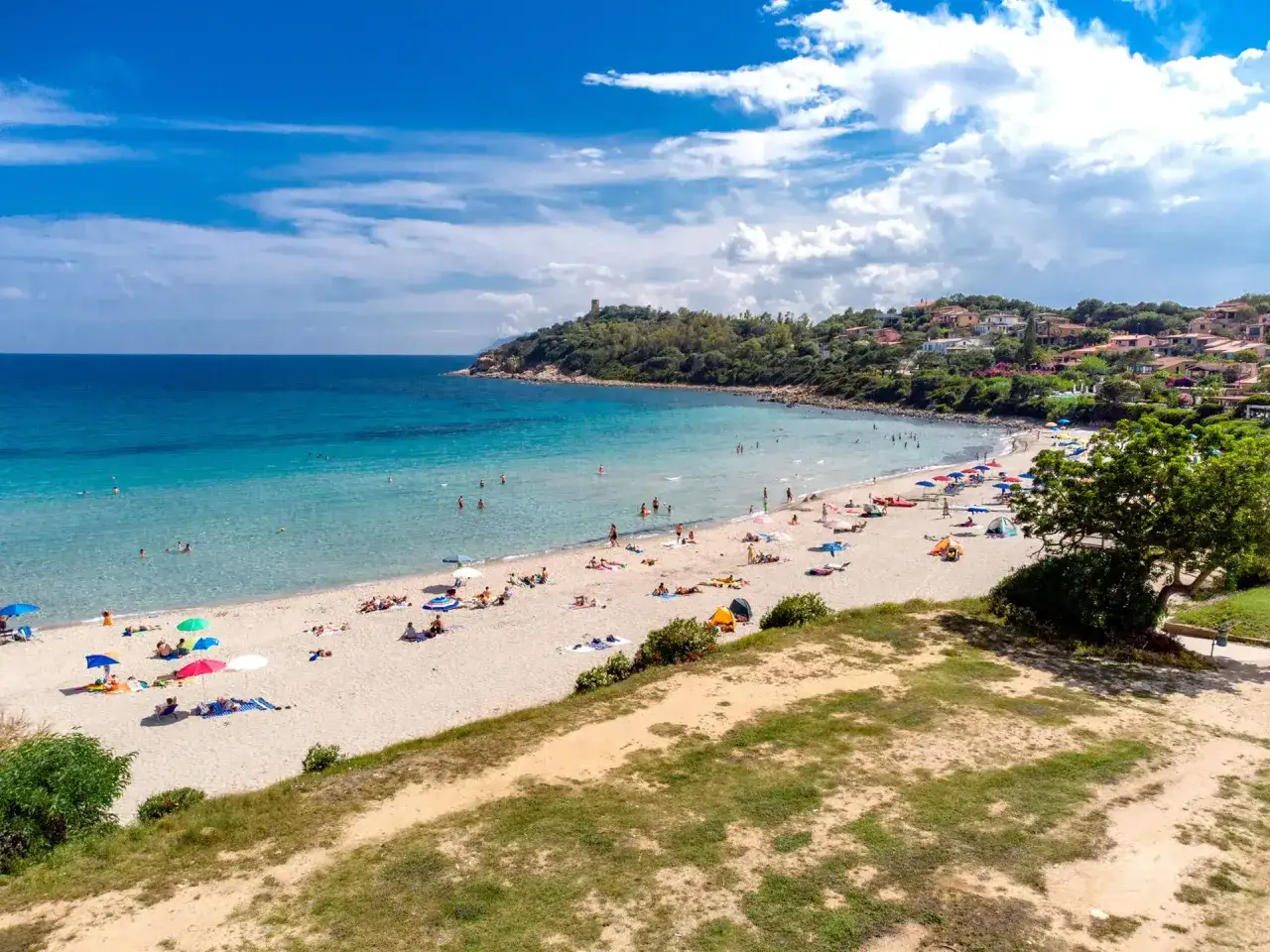 Spiaggia di Porto Frailis, con gente che prende il sole e nuota nelle acque cristalline sotto un cielo azzurro con nuvole bianche.