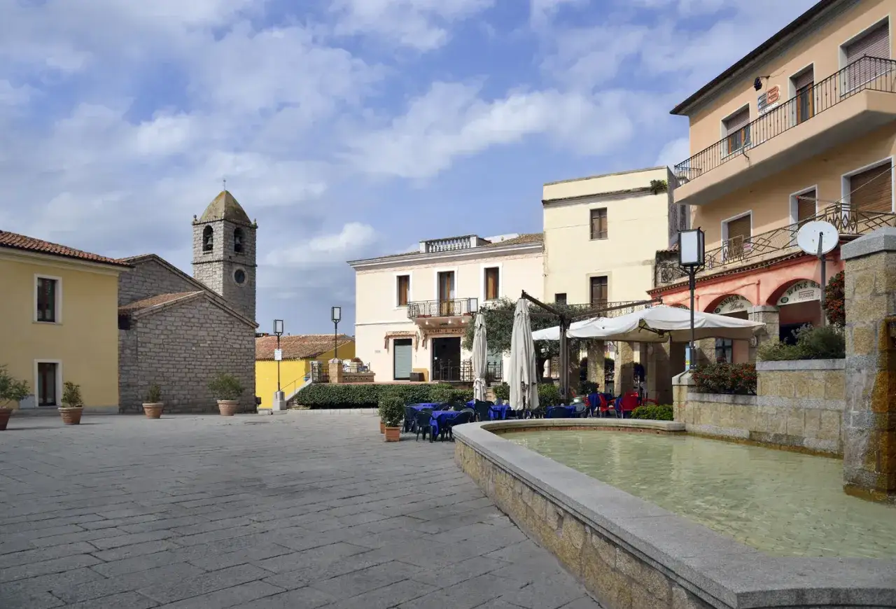 Piazza con fontana e tavolini all'aperto nel centro storico di Arzachena. Campanile in pietra sullo sfondo.