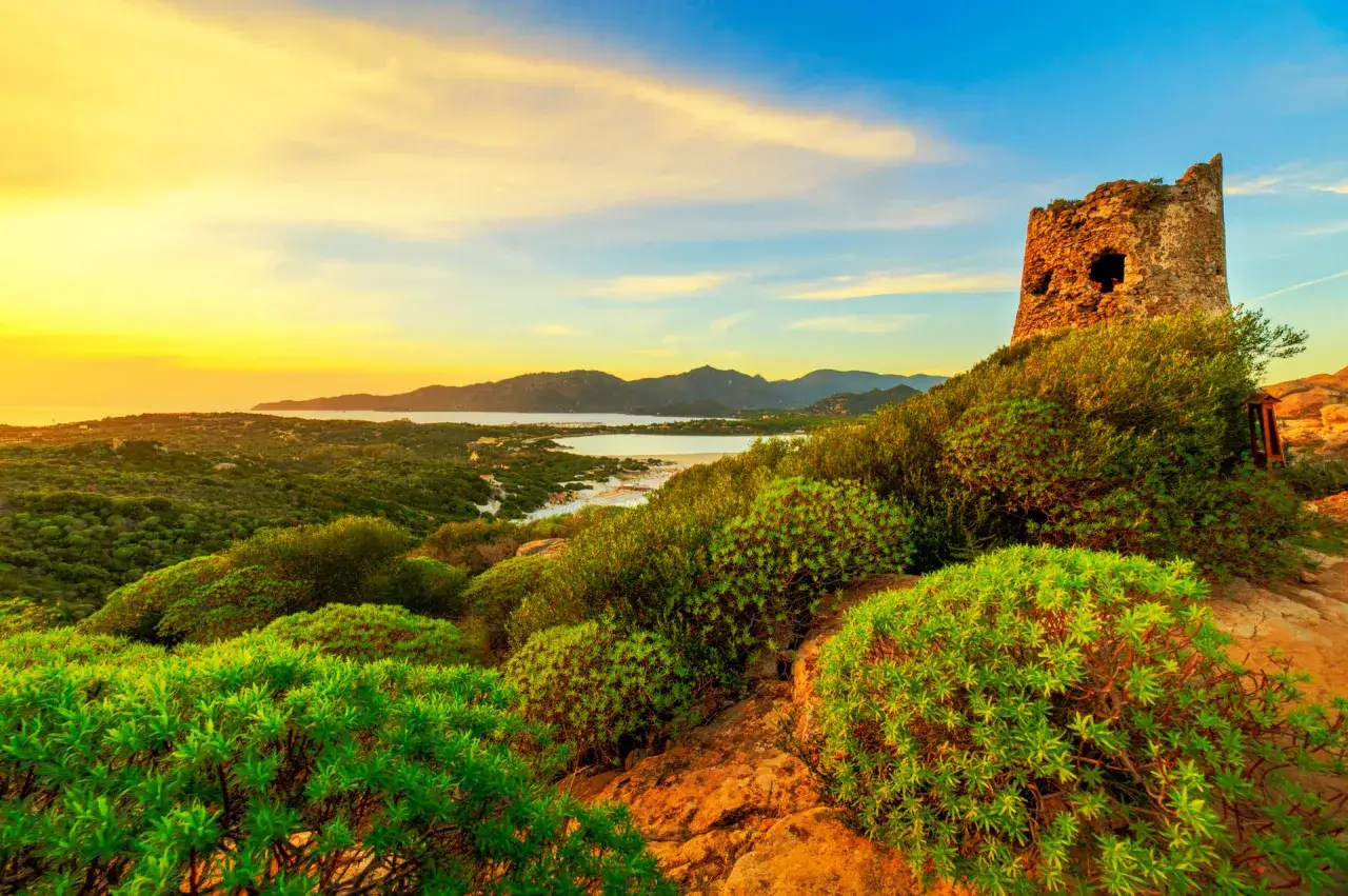 Torre di Porto Giunco, un'antica sentinella di pietra, domina un paesaggio di macchia mediterranea al tramonto, con il mare e le montagne sullo sfondo.