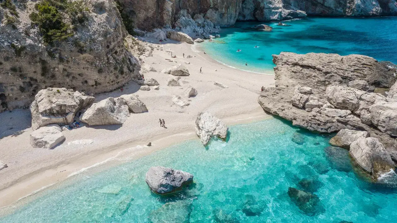 Spiaggia bianca e acque cristalline del mare della Sardegna, con scogliere rocciose e qualche persona che si gode il sole e il mare.