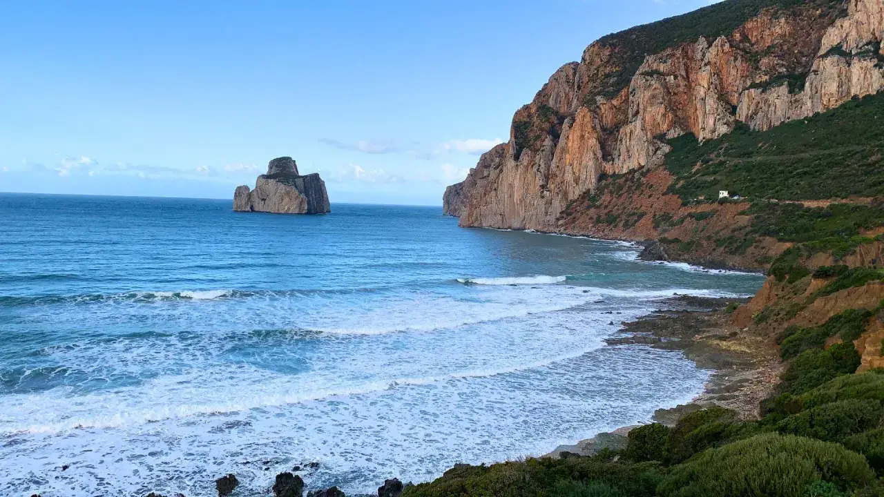 Scogliera rossa e mare blu a Nebida, Sardegna. Faraglioni imponenti emergono dall'acqua, mentre onde bianche lambiscono la costa rocciosa.
