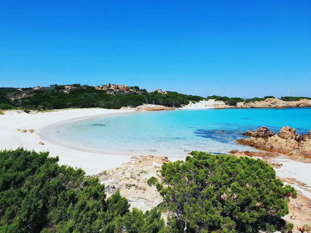 Spiaggia di Budoni con sabbia bianca e acque cristalline turchesi, incorniciata da rocce rosse e macchia mediterranea.