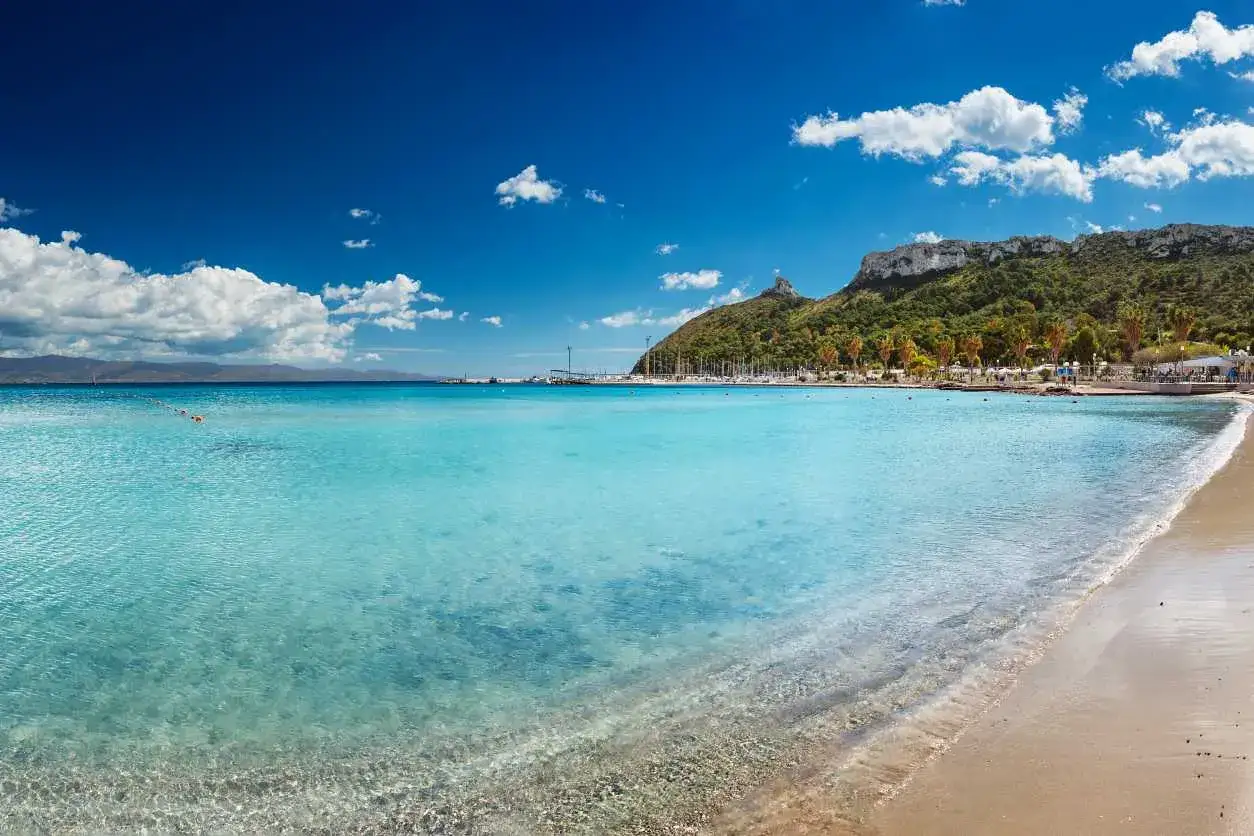 Acque cristalline e sabbia dorata sulle spiagge di Cagliari, con il promontorio verdeggiante e il porto turistico sullo sfondo.