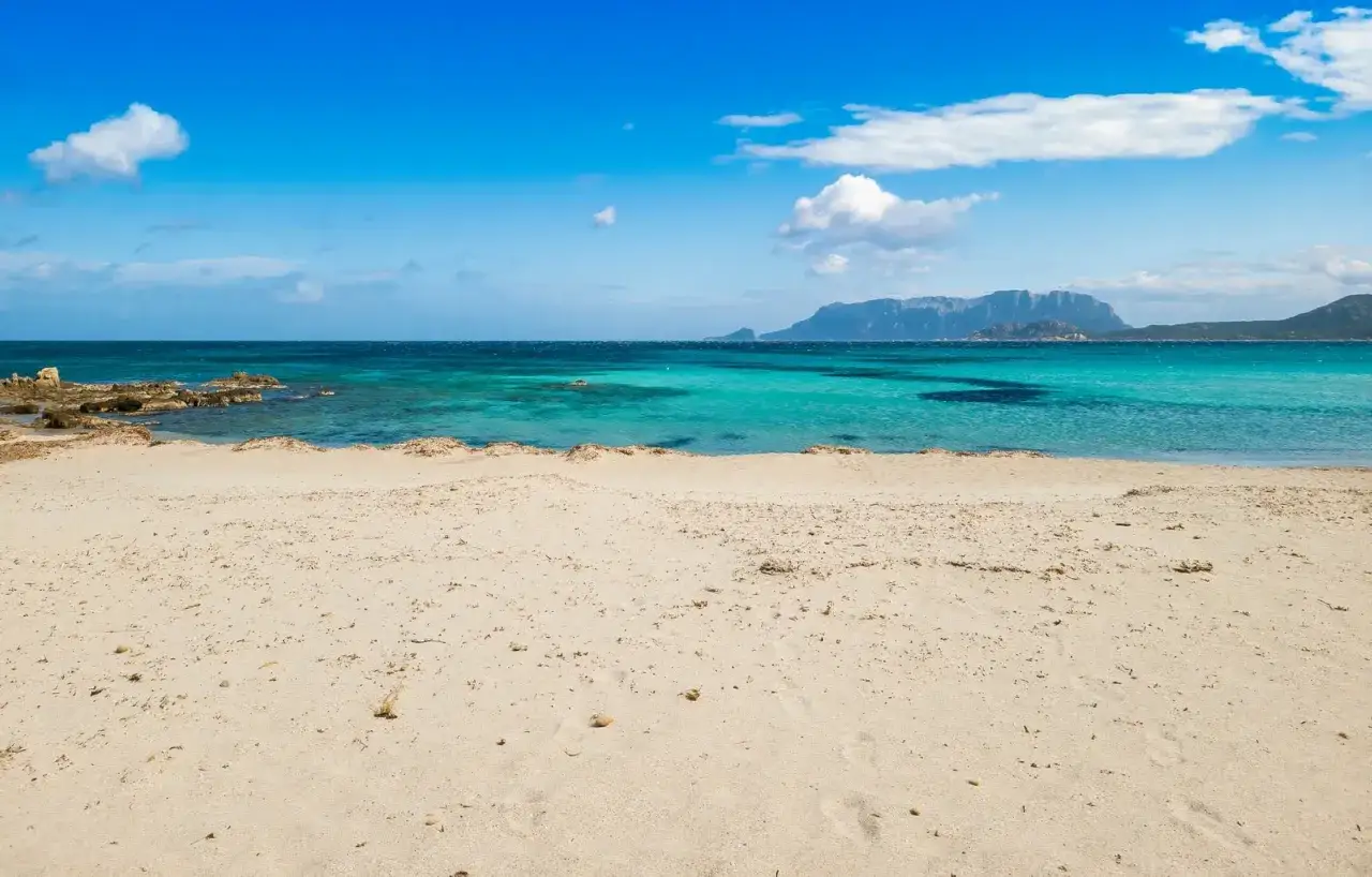 Spiaggia Pittulongu con sabbia dorata, mare turchese e montagne all'orizzonte sotto un cielo azzurro con nuvole bianche.