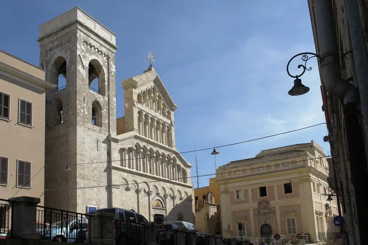 La piazza del palazzo a Cagliari, con la torre campanaria e la facciata della cattedrale sotto un cielo azzurro.
