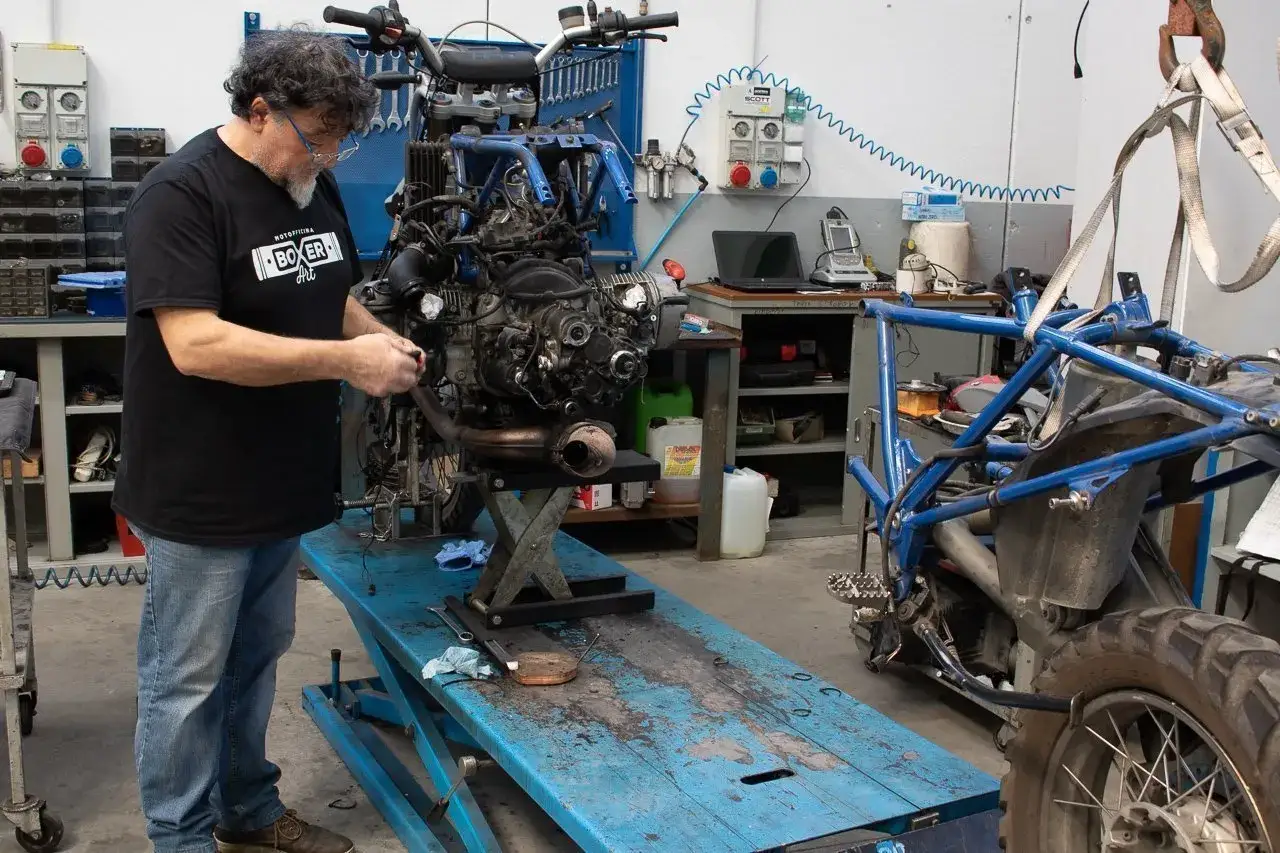 Meccanico lavora su motore moto, forse per come spurgare pompa frizione. Officina con attrezzi e telai moto.